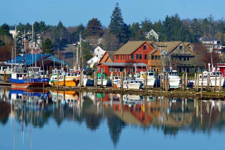 A colorful image of Ilwaco as seen from the lake. The brightly colored boats contrasted with the red buildings and blue water gives the picture an idyllic feeling.