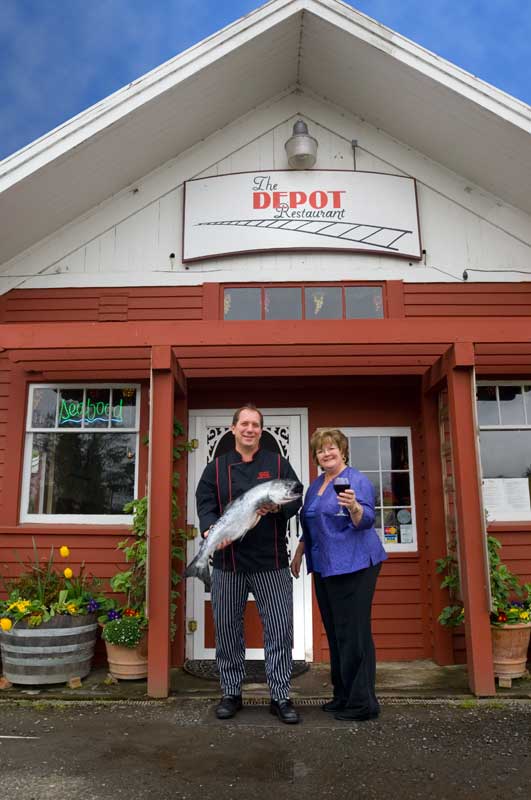 An image of The Depot Resteraunt with its owner, Bruce, standing out front with his wife, proudly holding the catch of the day - a bright, hefty salmon.