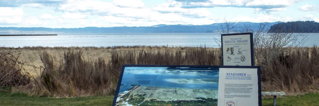 An image of a tourist observation point with a plaque in front that reads, "Remember, there's the face of a family behind every farmed oyster." overlooking the inlet with blue mountains in the background.