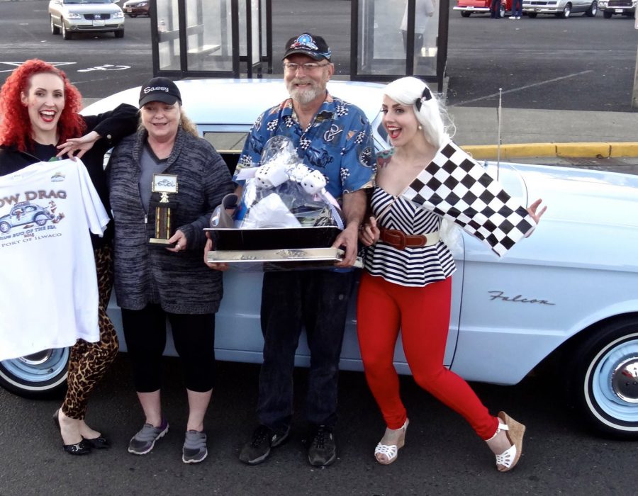 Image of locals posing in front of their classic car, having won a competition and holding the trophy and gift basket with pride.