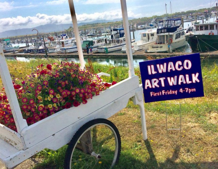 Image of a sunny, flower-filled white wheelbarrow next to a sign proclaiming the Ilwaco Art Walk event will be happening soon.