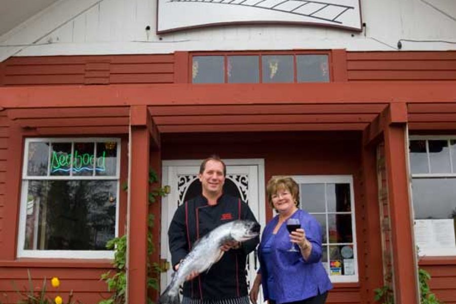 An image of The Depot Resteraunt with its owner, Bruce, standing out front with his wife, proudly holding the catch of the day - a bright, hefty salmon.