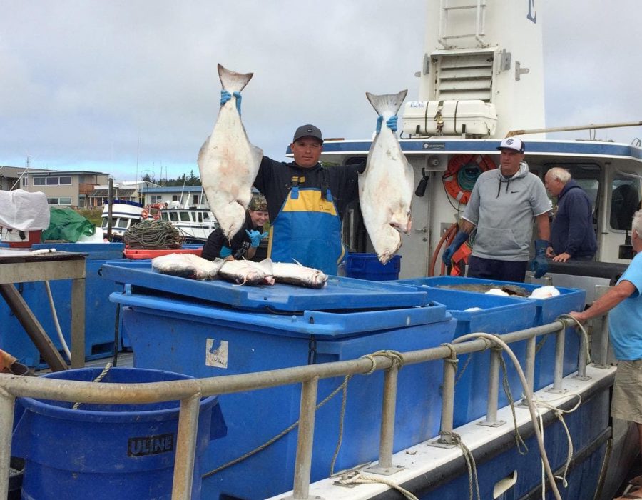 An image taken from the back of a large fishing boat where one worker holds up two impressively sized halibut, one in each hand, with pride.