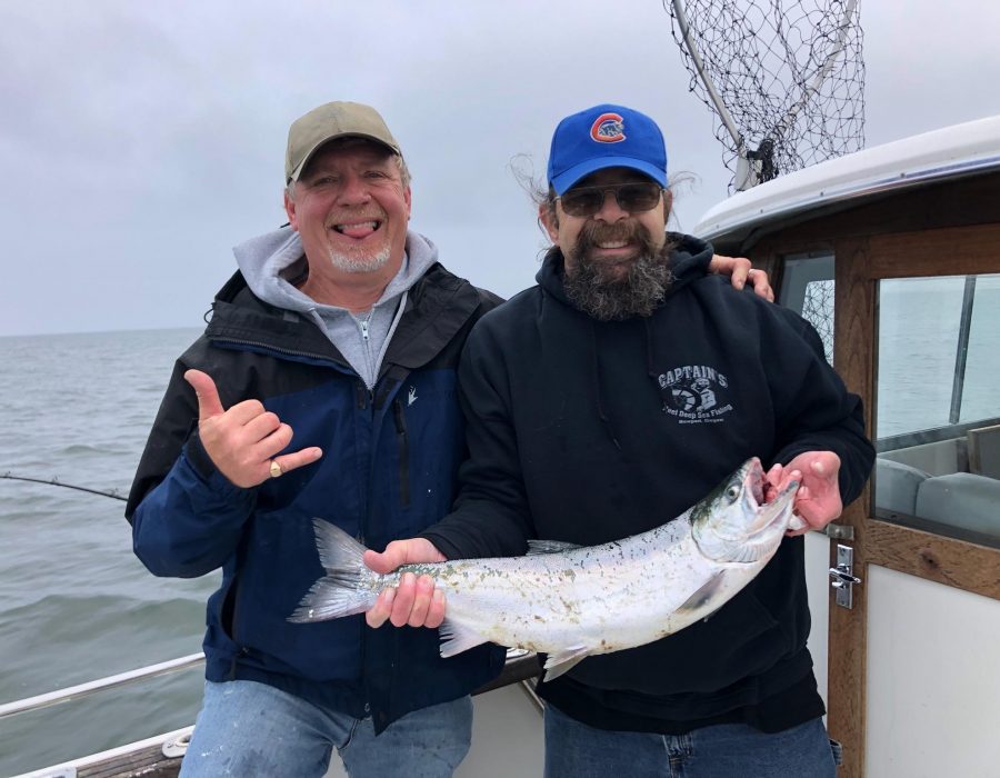 An image of two lucky locals out at sea, one of them holding an impressive salmon while their friend makes the hang loose shaka sign with his hand.