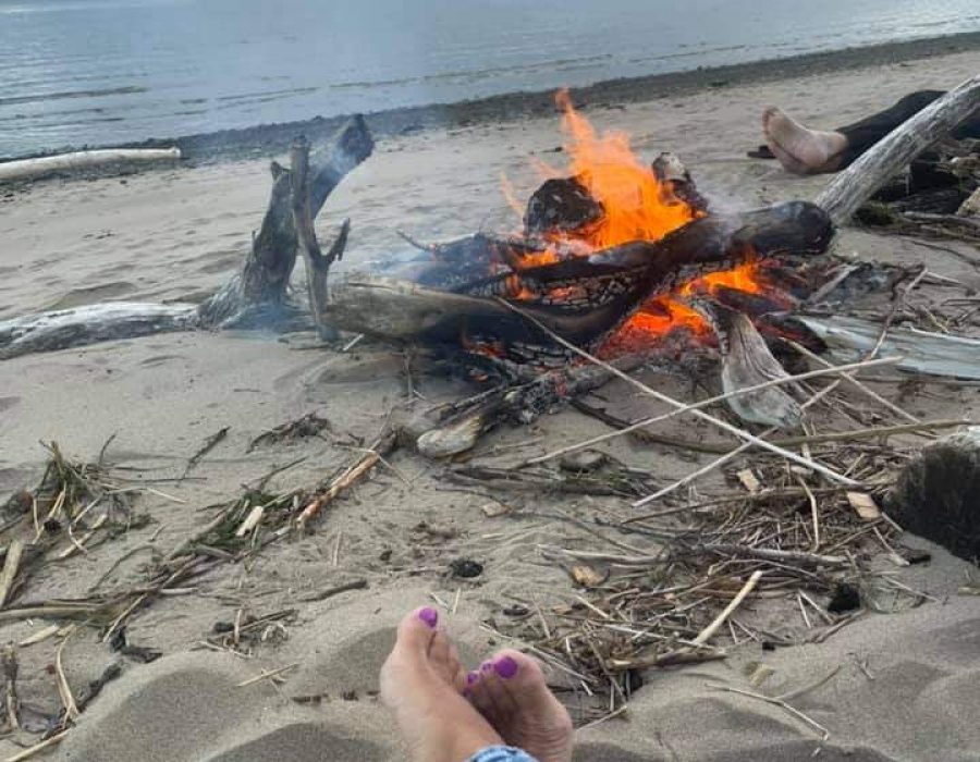 An image of a cozy fire built directly on the beach. A person's legs can be seen from the Point of View of the camera, as if you were the one relaxing there, watching the tide.