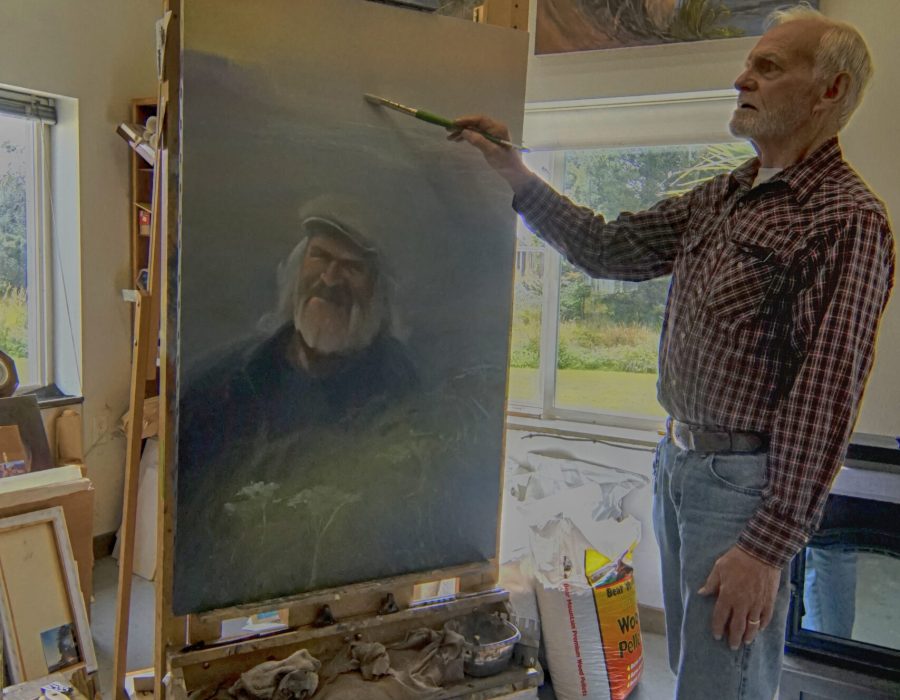 An image of Wallymann, a local Tokeland painter as he poses next to one of his paintings, pretending to paint on it. The painting is of and old man who looks like a sea captain.